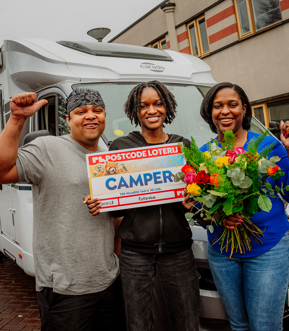 Drie mensen glimlachen voor een camper. Ze houden een cheque van de Postcode Loterij vast. Een vrouw heeft een boeket bloemen in haar hand.