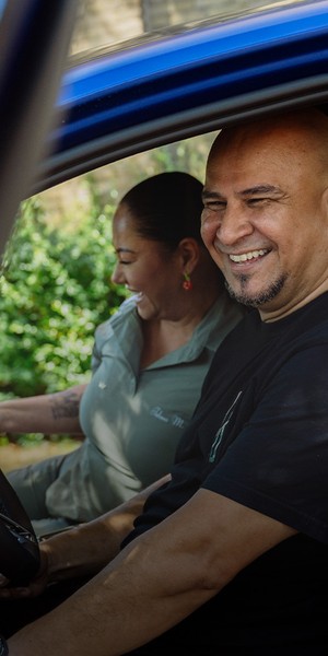 Een lachende man en vrouw zitten samen in een blauwe auto, kijkend naar iets buiten beeld. De man zit achter het stuur.