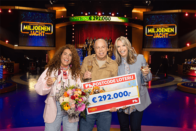 rie mensen staan lachend op een verlicht tv-podium tijdens de Postcode Loterij Miljoenenjacht. De persoon in het midden houdt een grote cheque van €292.000 vast, terwijl alle drie een glas champagne en een boeket bloemen vasthouden. Op de achtergrond zijn schermen met het logo van Miljoenenjacht en publiek zichtbaar.